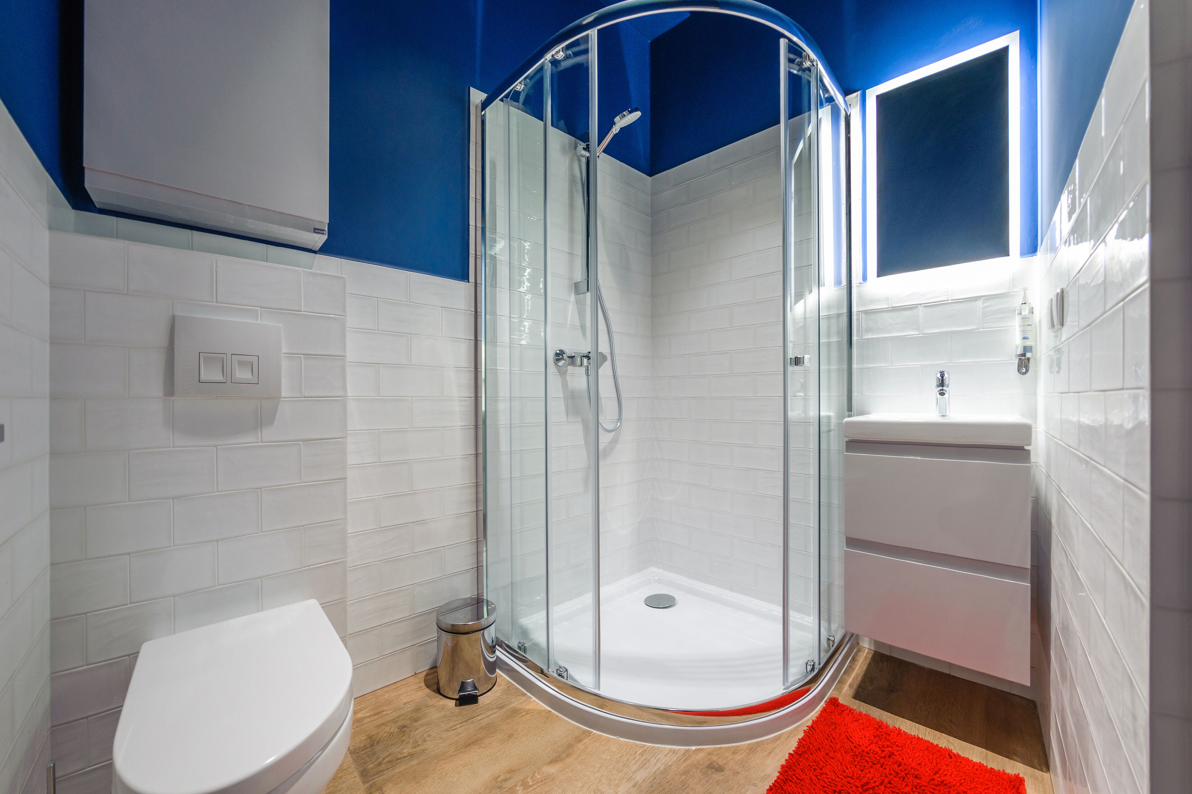 A modern bathroom with a glass-enclosed shower, white tiles, a white toilet, and a red rug.