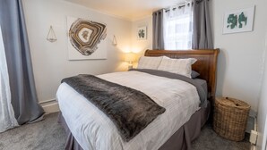 A neatly made bed with a grey and white duvet, a wooden headboard, and a grey furry throw blanket. There is a wicker basket beside the bed and a framed abstract painting on the wall. - Downtown 1 Bedroom Charmer - Walk, Bike, Visit, Enjoy!! (Rapid City)