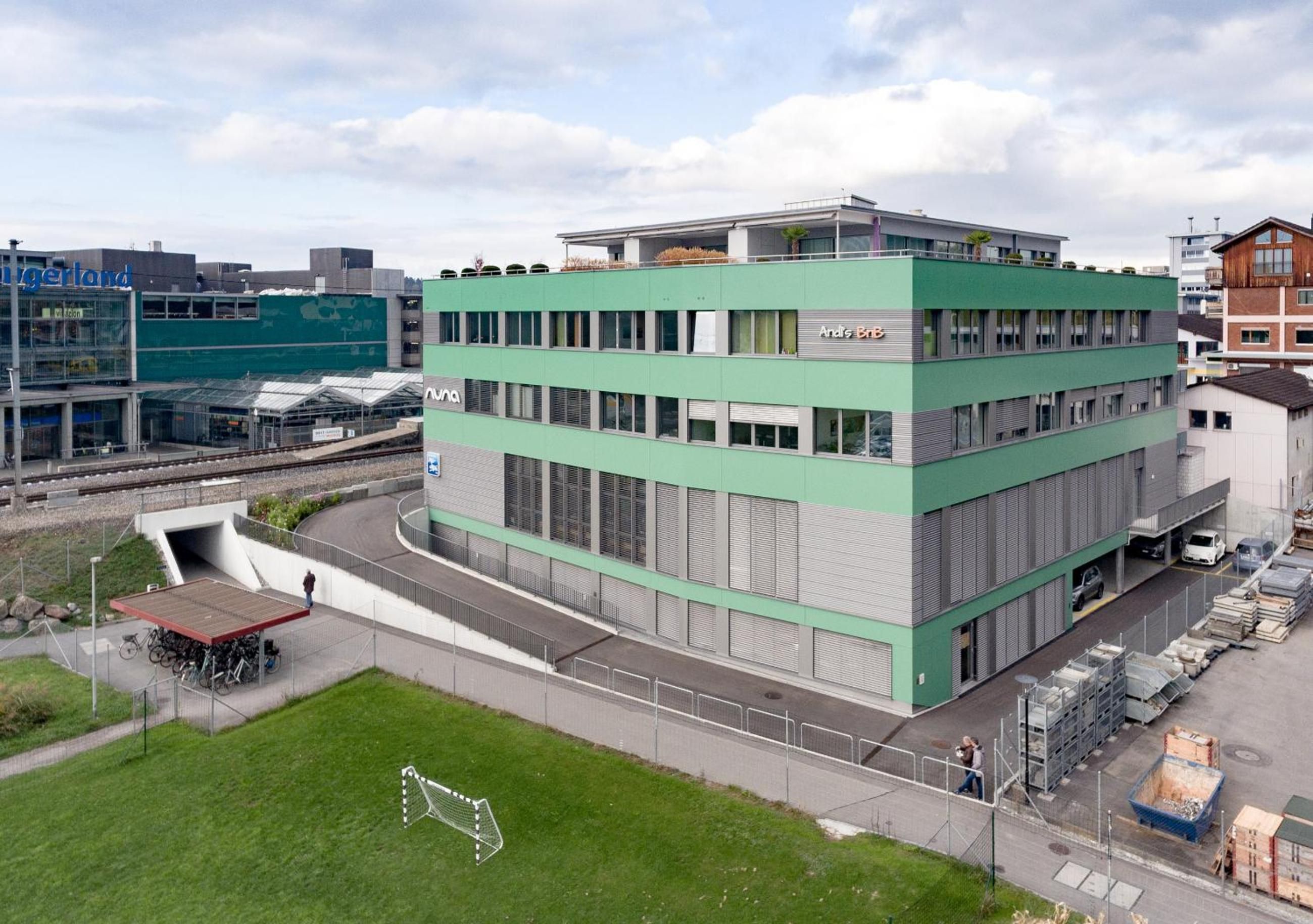 Ein mehrstöckiges Gebäude mit grüner Fassade, einem überdachten Eingang und einem Parkplatz mit Autos und Baumaterialien.