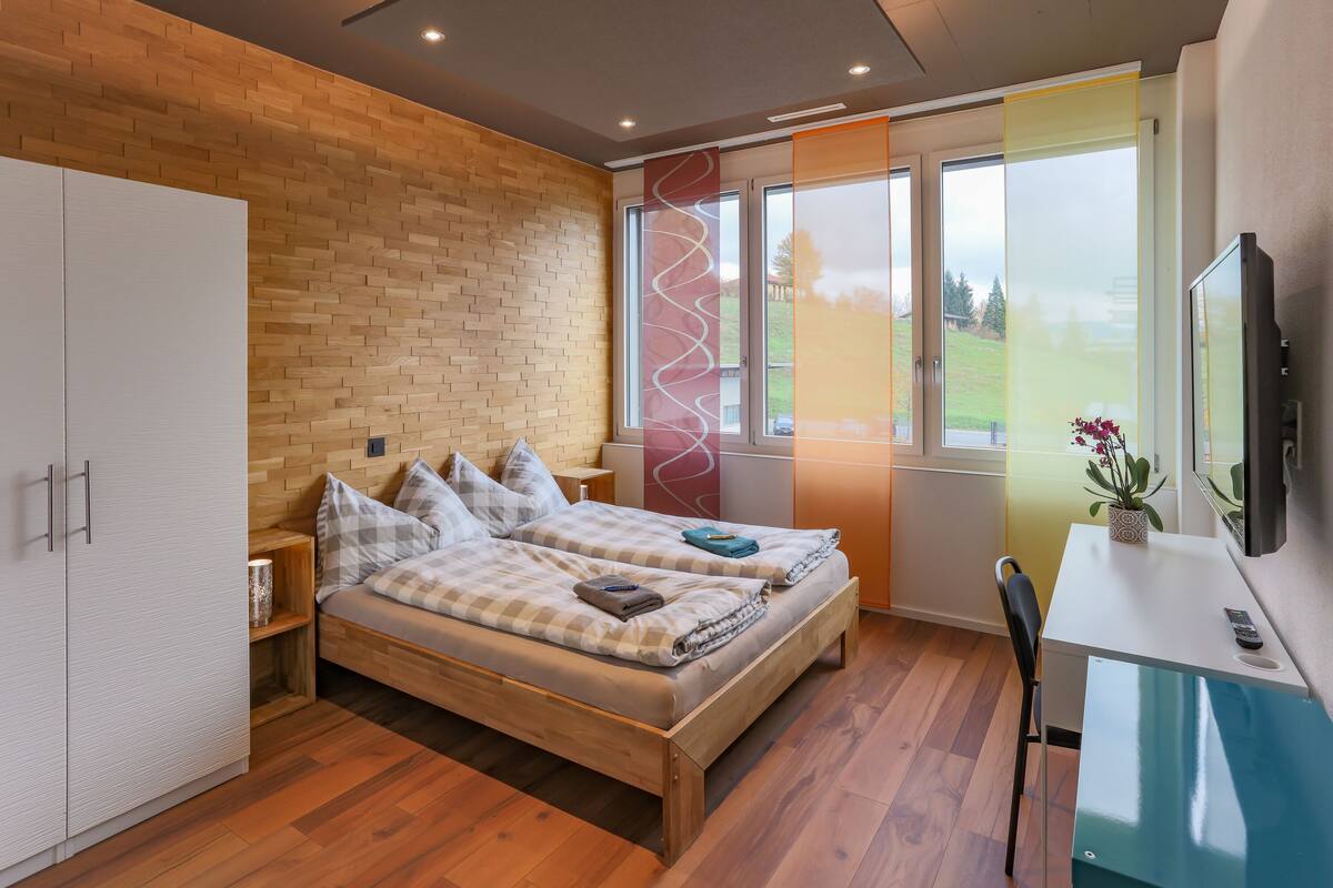 A bedroom with a wooden bed, a desk, a TV, and a window with a view of the outdoors.
