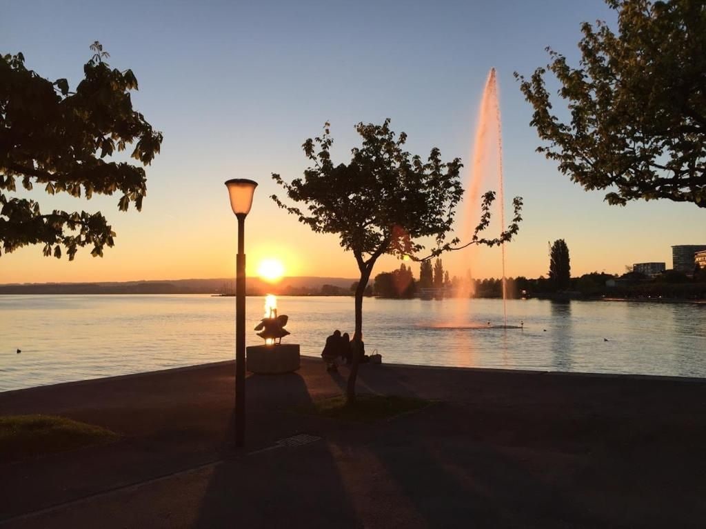 Vista do p ôr do sol com uma fonte, um poste de luz e um banco à beira de um lago.
