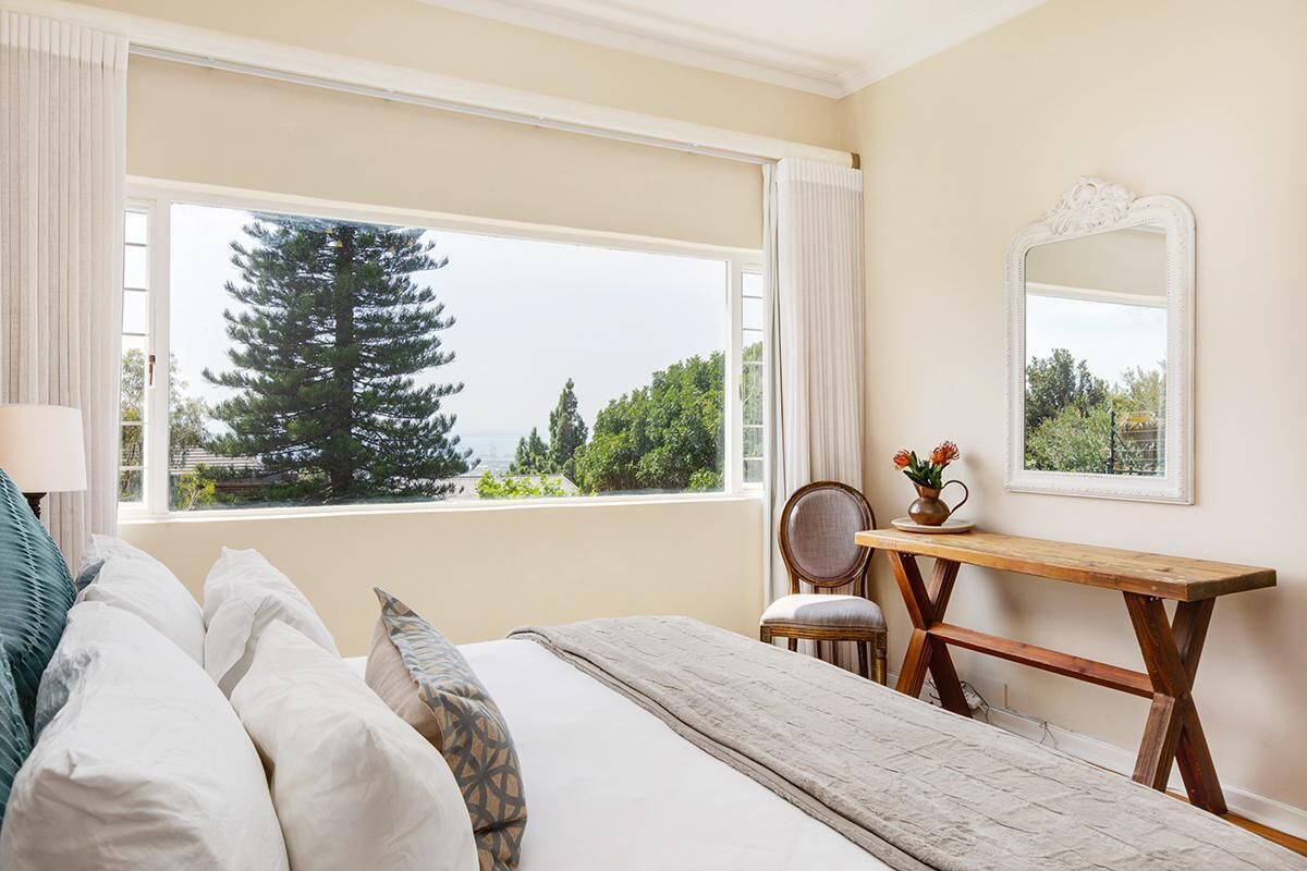 A bedroom with a large window, a bed with pillows, a wooden desk, a chair, and a mirror on the wall.