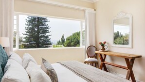 Ein Schlafzimmer mit großem Fenster, einem Bett mit Kissen, einem Holztisch, einem Stuhl und einem Spiegel an der Wand.