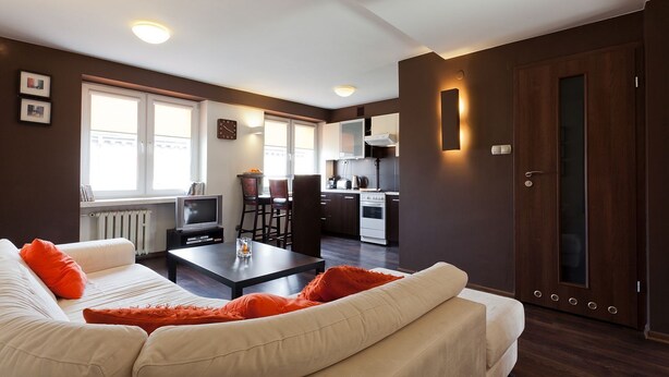 Una sala de estar moderna con un sofá, una mesa baja y un televisor. Se observa una zona de cocina con estufa y alacenas.