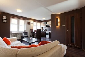 Una sala de estar moderna con un sofá, una mesa baja y un televisor. Se observa una zona de cocina con estufa y alacenas.