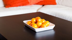 Un sofá blanco con almohadones naranjas, una mesa blanca con un cuenco de naranjas, y una pared adornada con cuadros enmarcados.