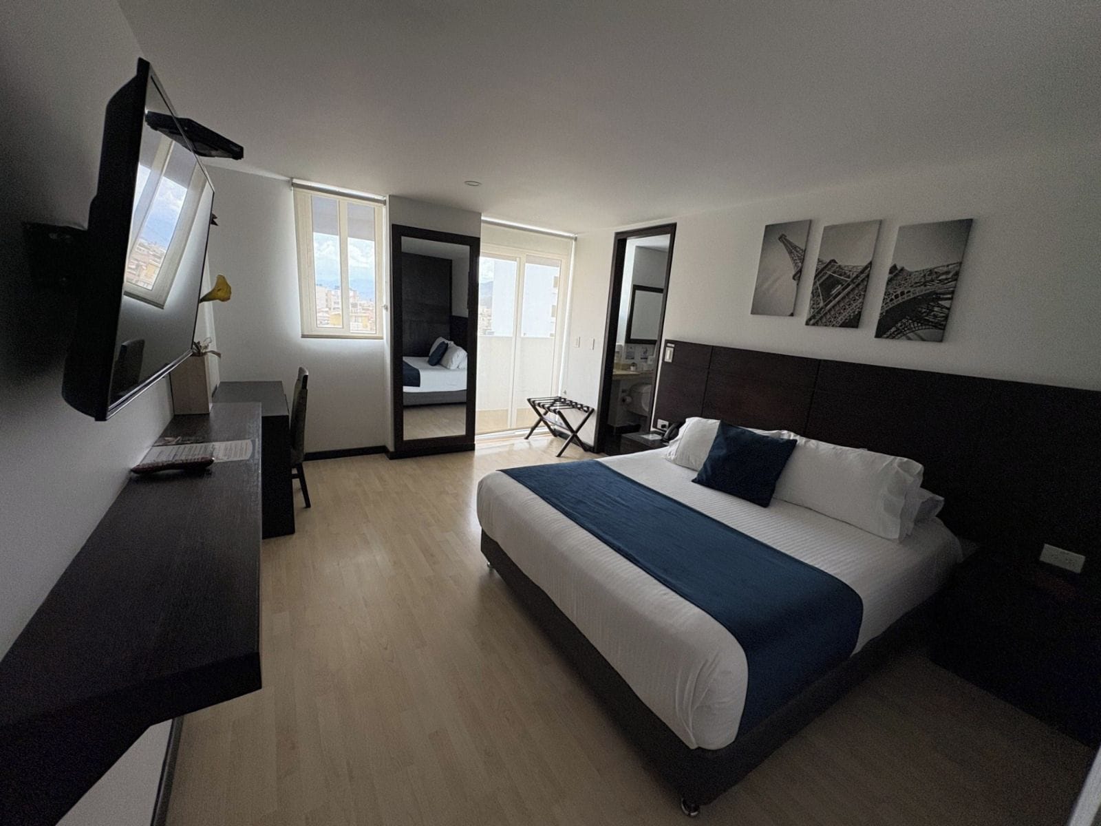 Una habitación de hotel con cama, escritorio, silla, televisor y obras de arte en la pared.