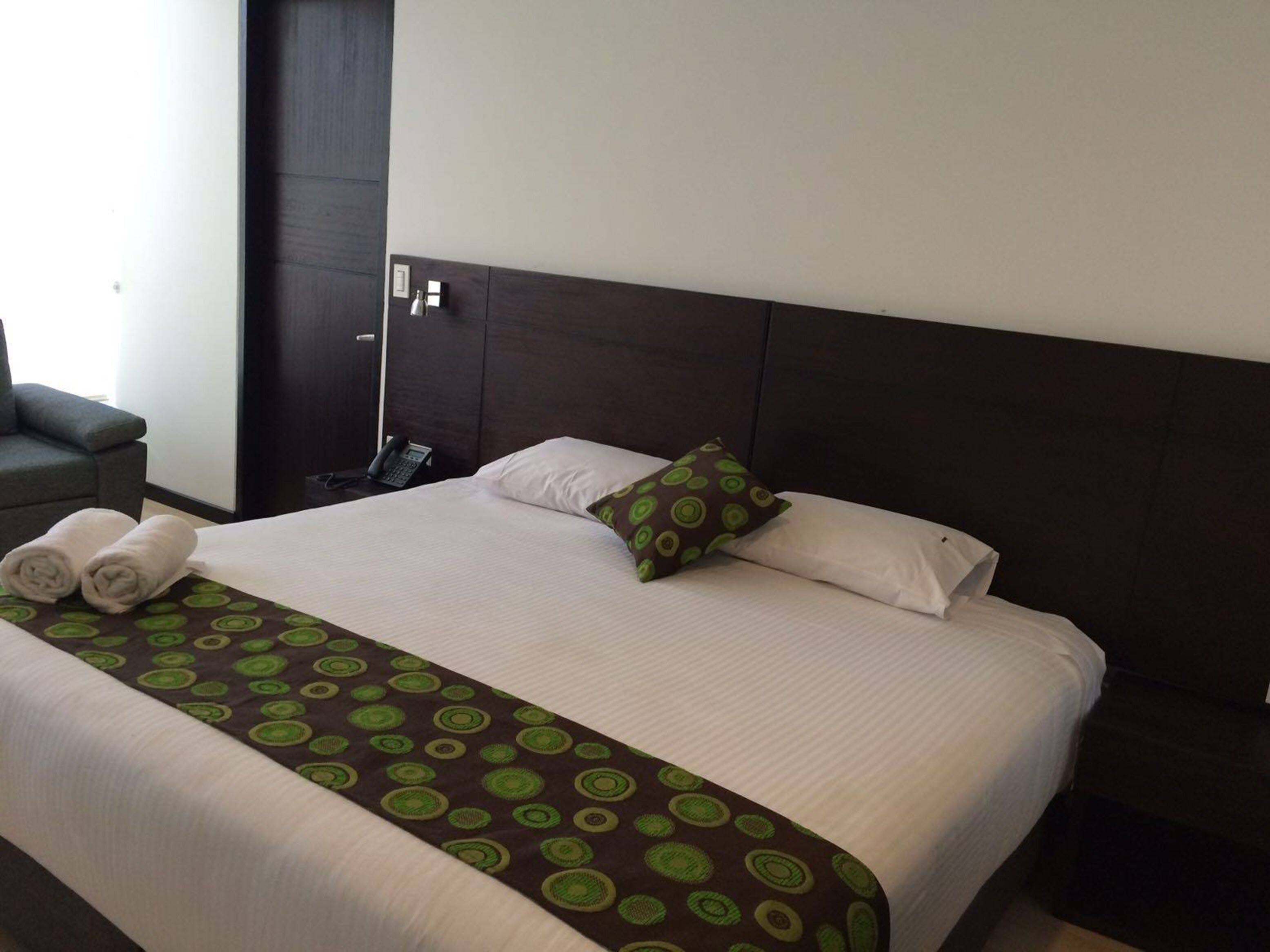 A neatly made bed with a patterned bedspread, pillows, and rolled towels on a dark headboard.