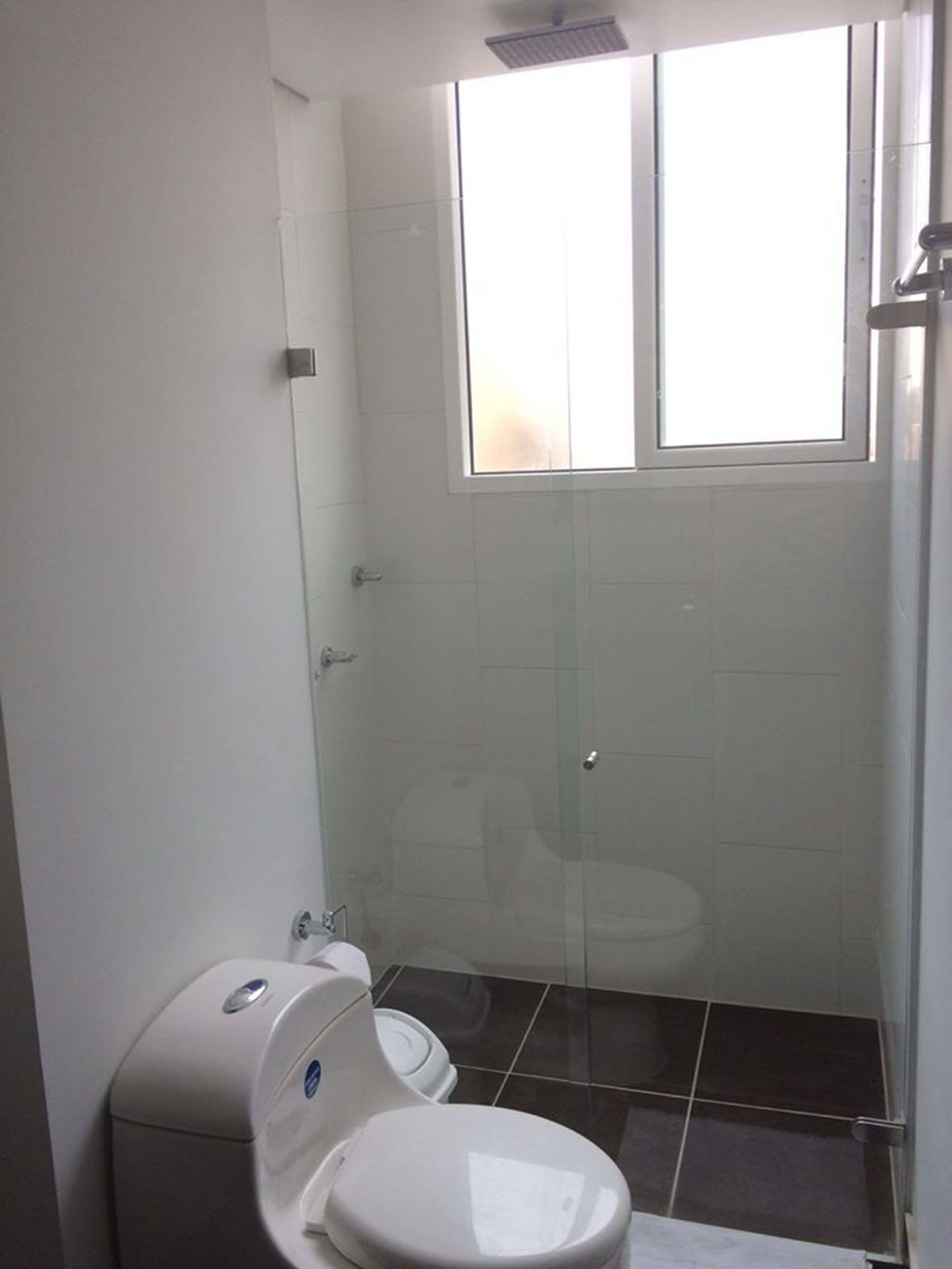 Une petite salle de bain compacte, dotée de toilettes, d’une douche à parois de verre et d’une fenêtre laissant entrer la lumière naturelle.