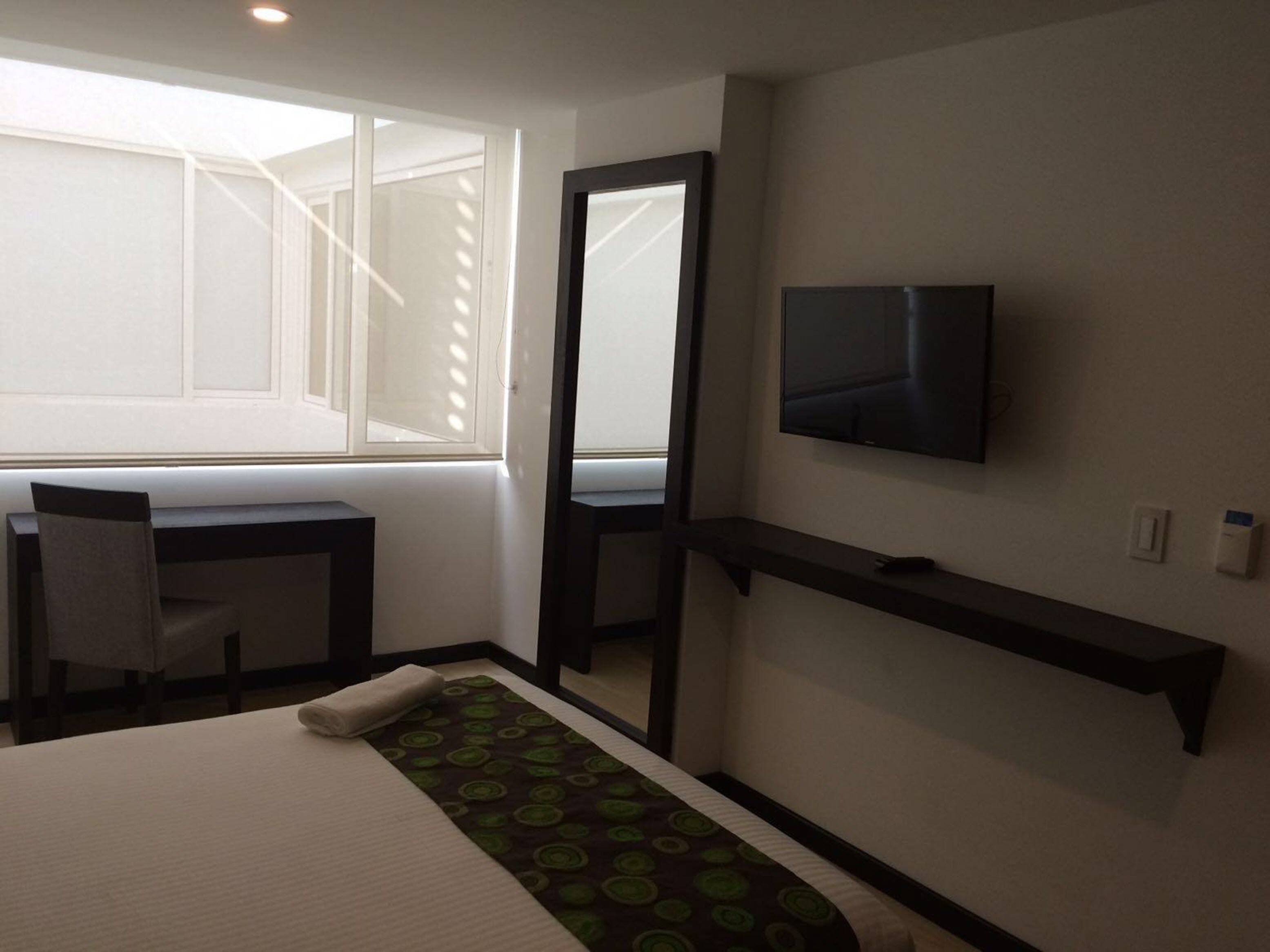A hotel room with a bed, a desk, a chair, a TV mounted on the wall, and a window with blinds.