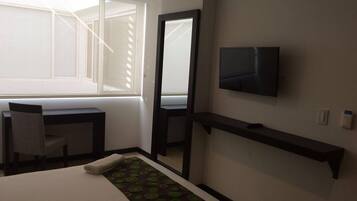 A hotel room with a bed, a desk, a chair, a TV mounted on the wall, and a window with blinds.