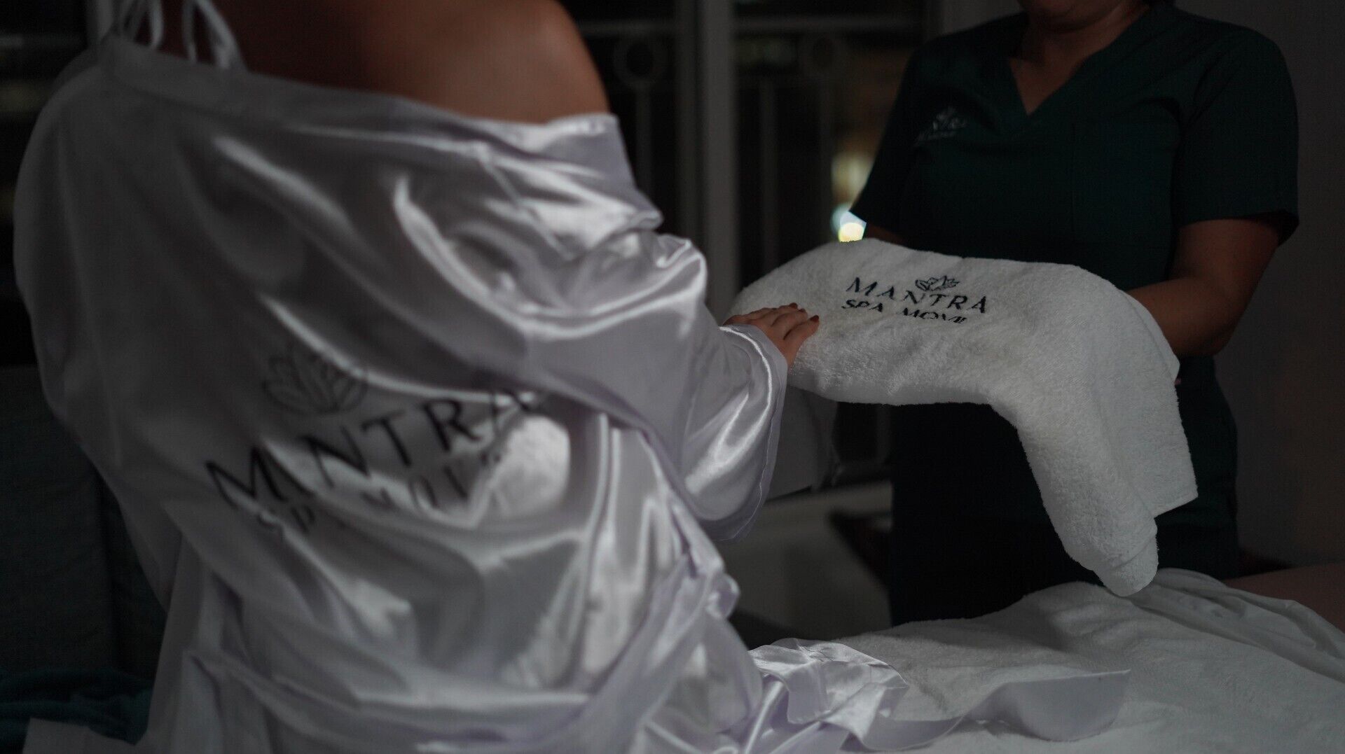 A person in a white robe with 'MANTRA SPA MOVEMENT' on the back, holding a white towel with the same text.