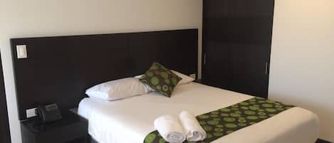Un lit bien fait, avec des draps blancs, un plaid à motifs verts et des serviettes enroulées.