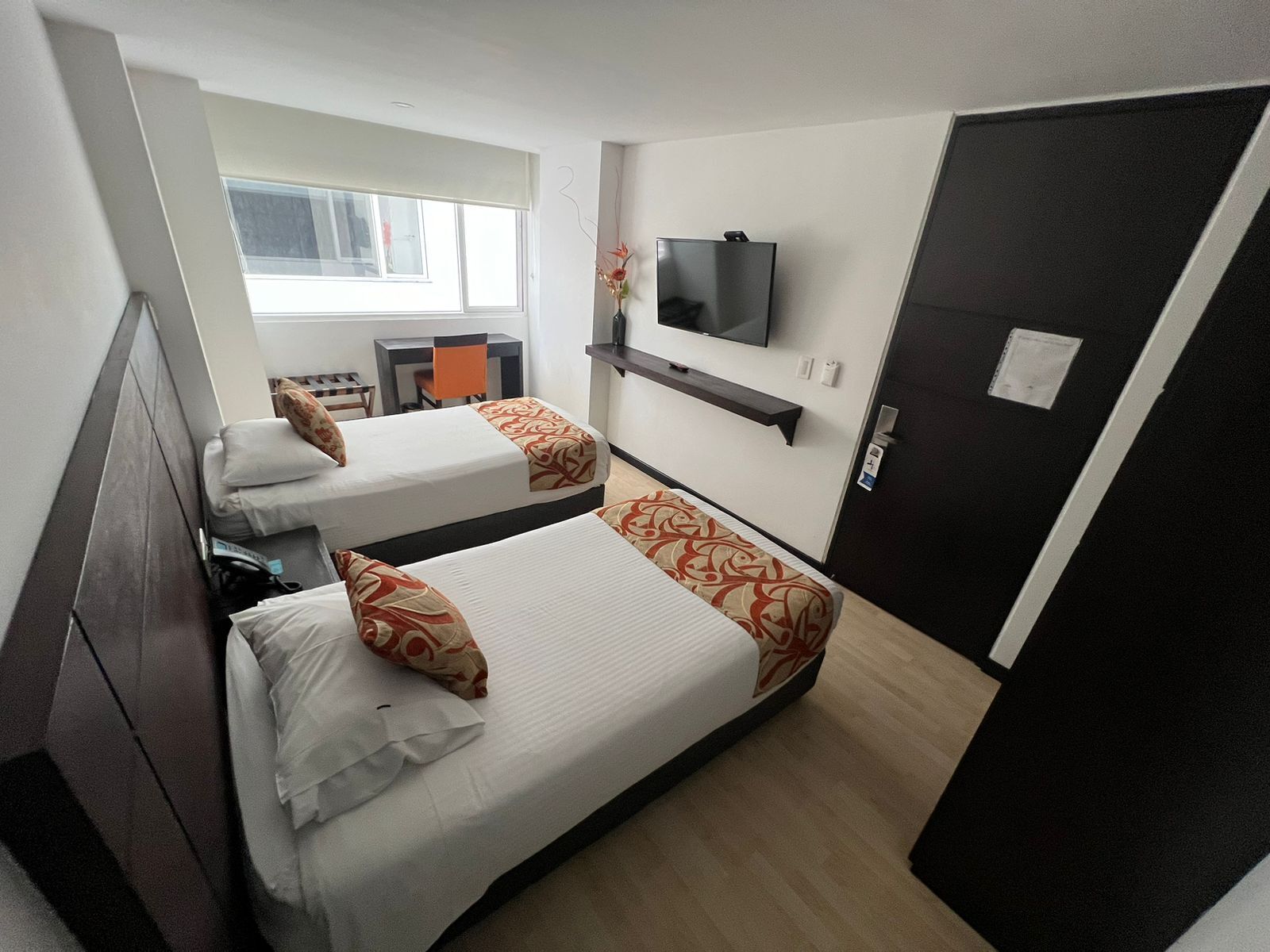 Een hotelkamer met twee bedden, een aan de muur bevestigde televisie, een bureau met een stoel, en een raam met jaloezieën.