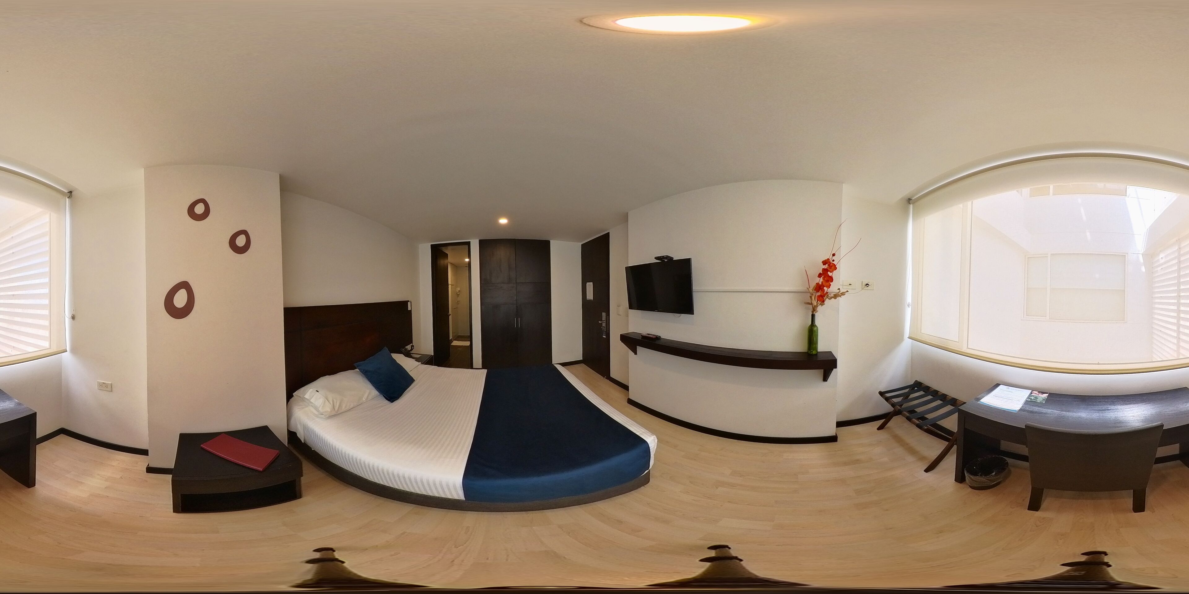 Habitación de hotel compacta con cama curva, escritorio pequeño, televisor y ventana con persianas.