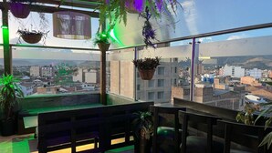 A rooftop terrace with a view of a cityscape, featuring a wooden bench, chairs, hanging planters, and a pergola structure.