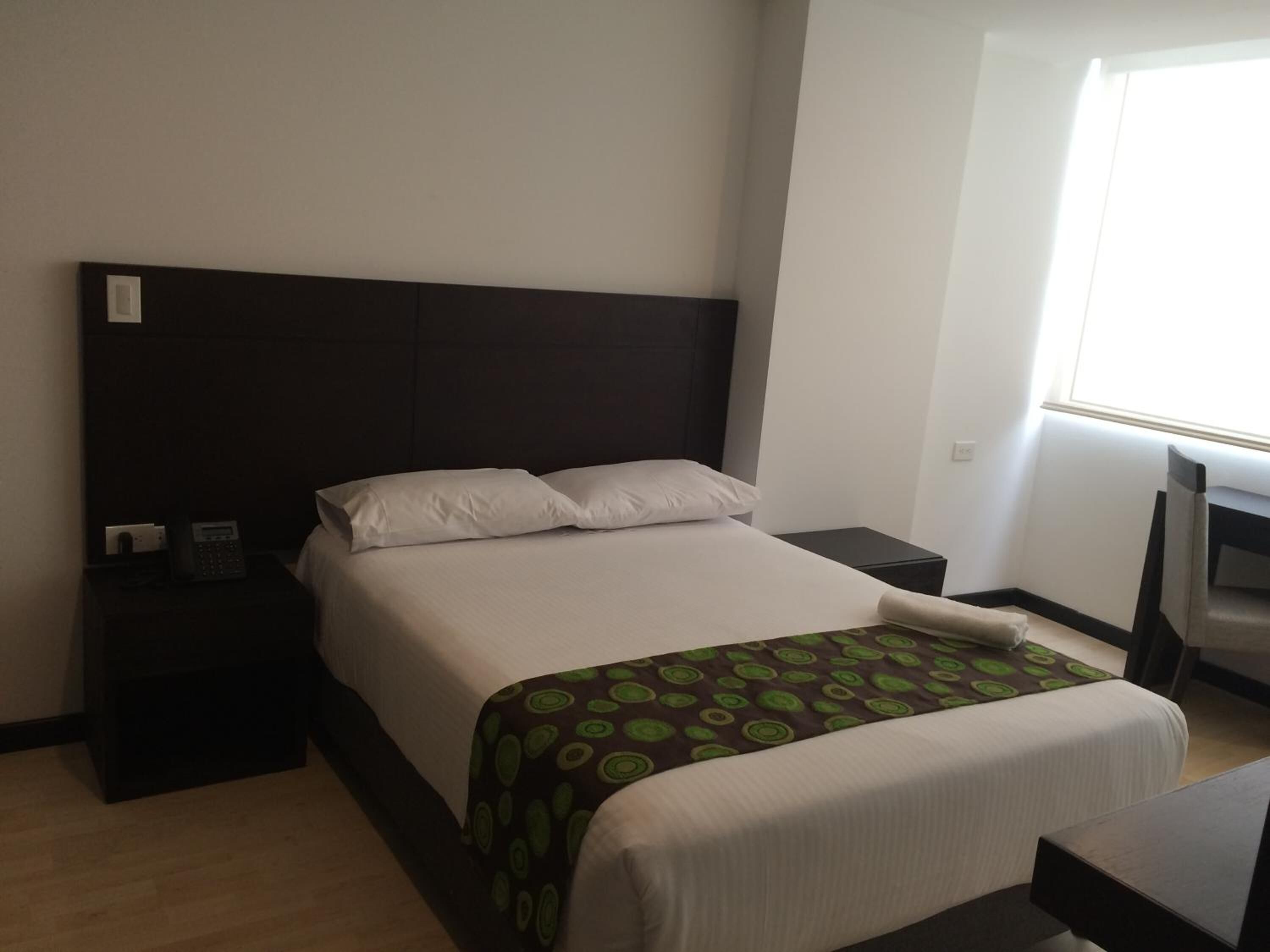 Une chambre d’hôtel avec un lit, une table de chevet avec un téléphone, une télévision et une fenêtre laissant entrer la lumière naturelle.