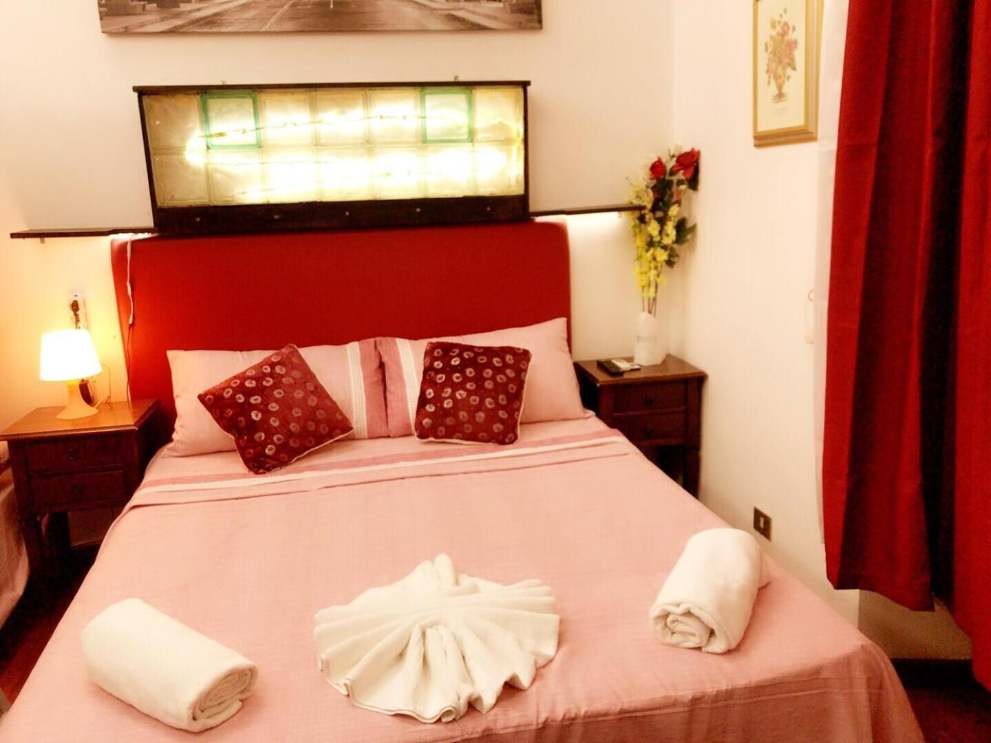 A hotel room with a bed, pillows, a nightstand, a lamp, a vase with flowers, and a painting on the wall.