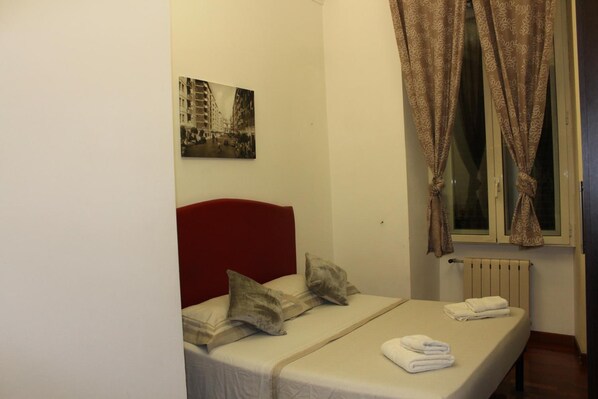 A small, neatly arranged bedroom with a single bed, a red headboard, a window with curtains, a radiator, and a wall-mounted picture.