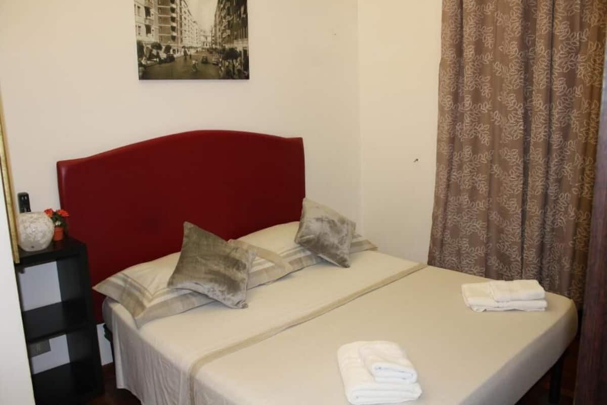 A hotel room with a red headboard, a bed with two pillows, a nightstand with a vase and flowers, and a folded towel on the bed.