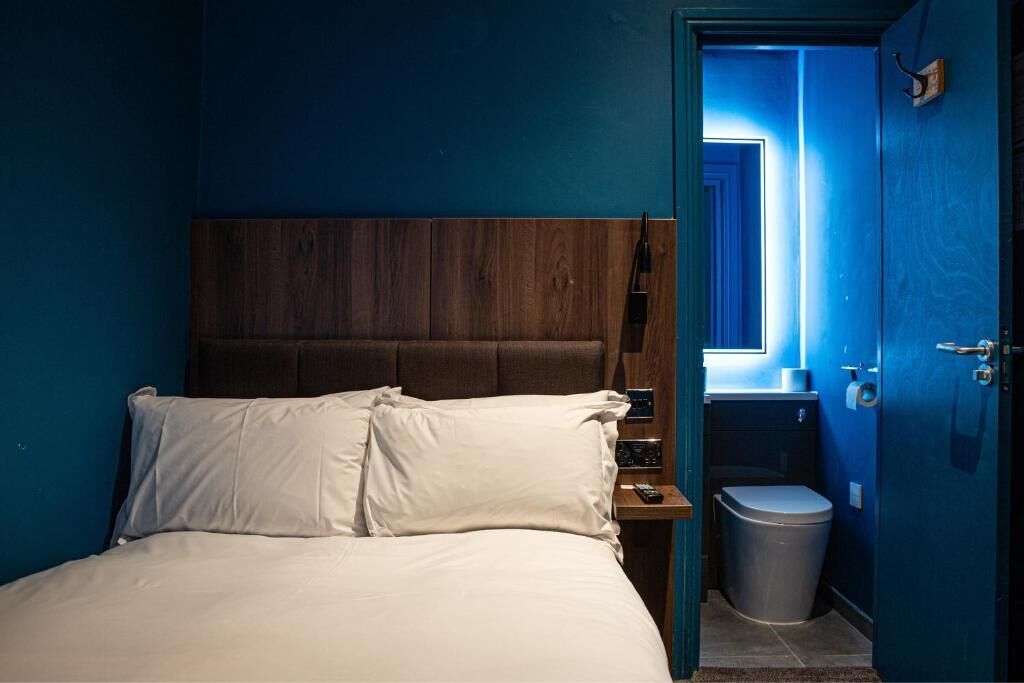 Un dormitorio con cabecera de madera, una cama individual con ropa blanca, una mesita de noche, un cubo de basura y un baño visible a través de una puerta abierta.