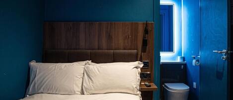 Un dormitorio con cabecera de madera, una cama individual con ropa blanca, una mesita de noche, un cubo de basura y un baño visible a través de una puerta abierta.