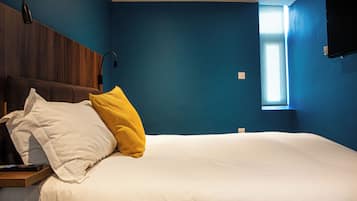 A bedroom with a bed, two pillows, a nightstand, a remote control, a wall-mounted TV, and a window.