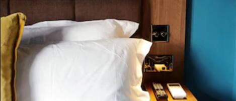 Un dormitorio con cabecera de madera, ropa de cama blanca, una laptop sobre la cama, una mesita de noche con una lámpara, y un buró con teléfono y control remoto.
