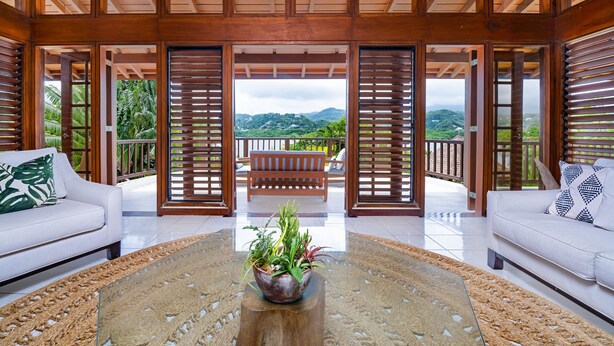 A spacious living area with a white sofa, a glass coffee table with a plant centerpiece, and a view of lush greenery and mountains through open wooden doors.