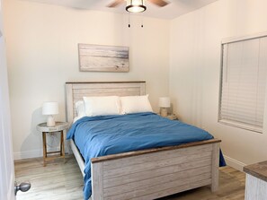 A bedroom with a wooden bed, two bedside tables with lamps, a ceiling fan, a painting on the wall, and a window with blinds.