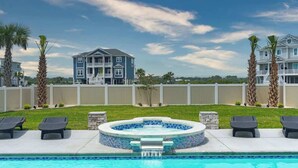 A pool area with lounge chairs, a hot tub, and a large building in the background. - Beach Please (Ocean Isle Beach)