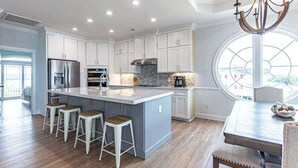 A modern kitchen with white cabinets, a central island with stools, a dining table with chairs, and a large window. - Beach Please (Ocean Isle Beach)