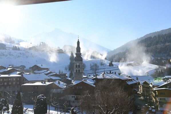 A snow-covered village with a prominent church tower, surrounded by residential buildings and a ski lift in the background.
