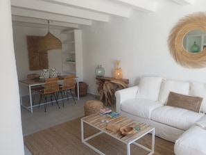 A modern living room with a white sofa, a wooden coffee table, wicker chairs, and a dining area with a white table and chairs.