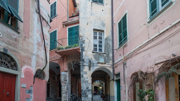 Un streetto vicolo fiancheggiato da edifici, caratterizzato da pareti in pietra, portali ad arco e una porta rossa a sinistra.