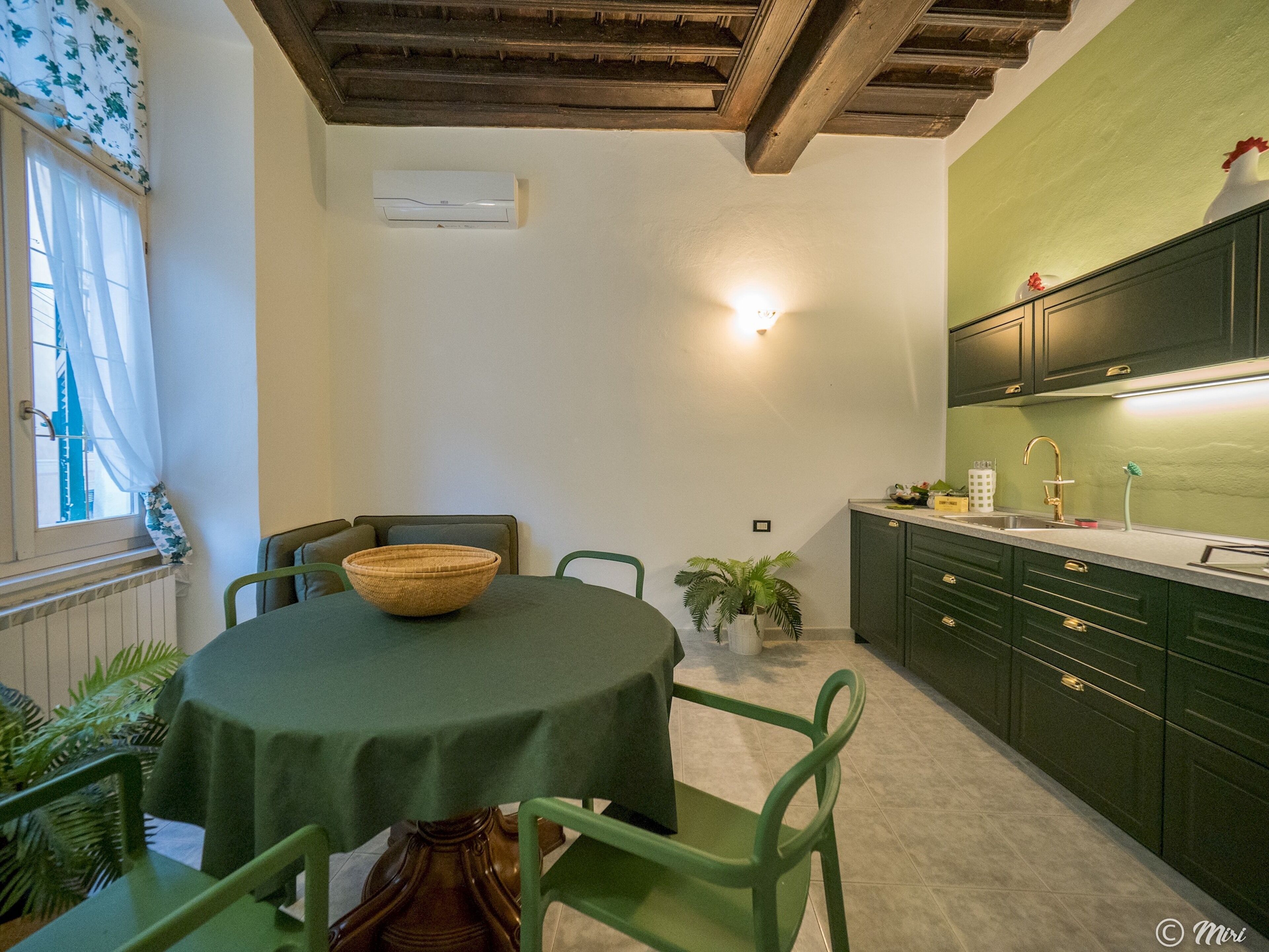 A dining area with a round table, chairs, a bowl, a potted plant, and a kitchen with green cabinets and a white countertop.