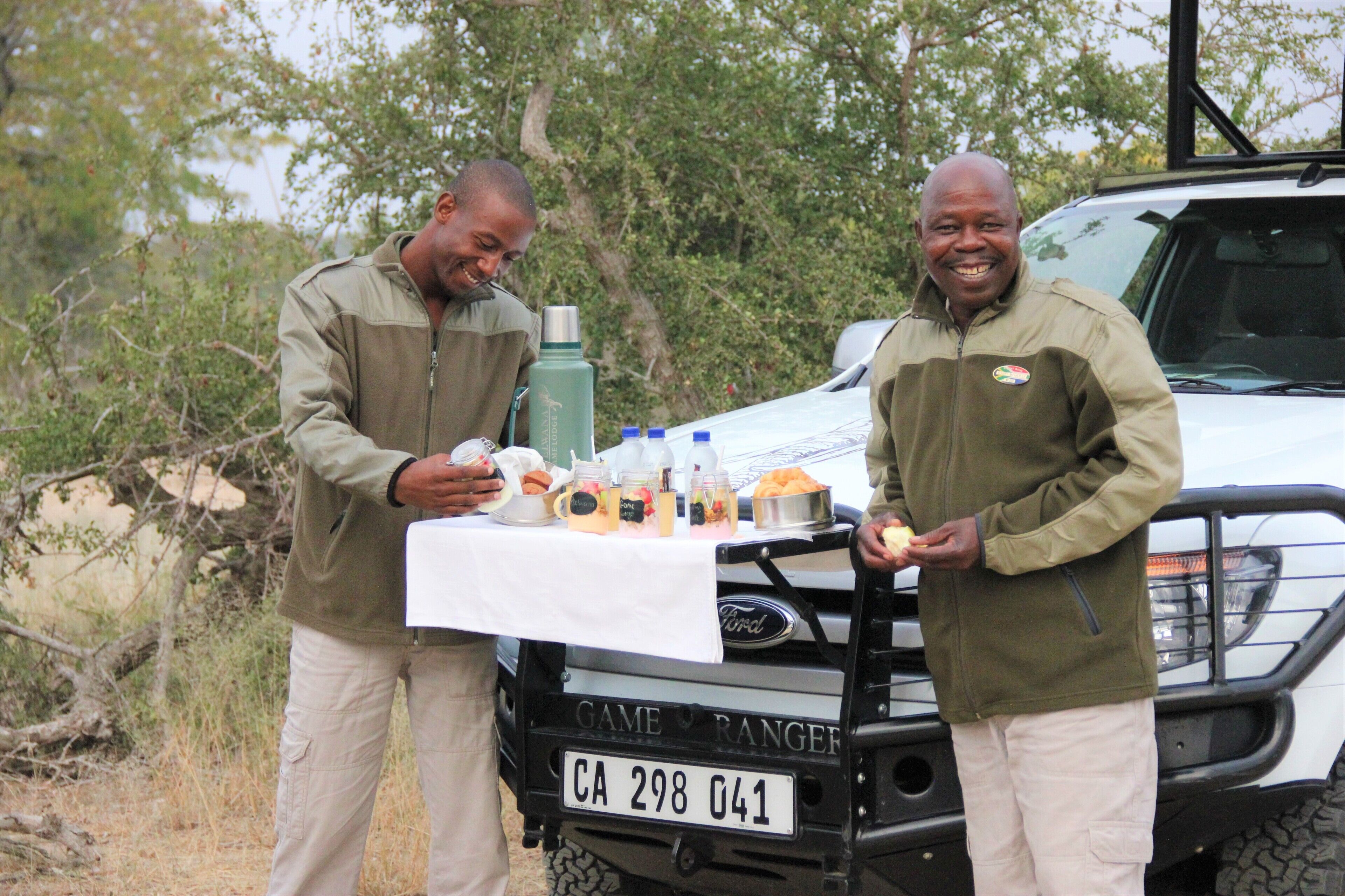 Two men standing by a table with food and drinks, in front of a vehicle with a license plate CA 298 041.