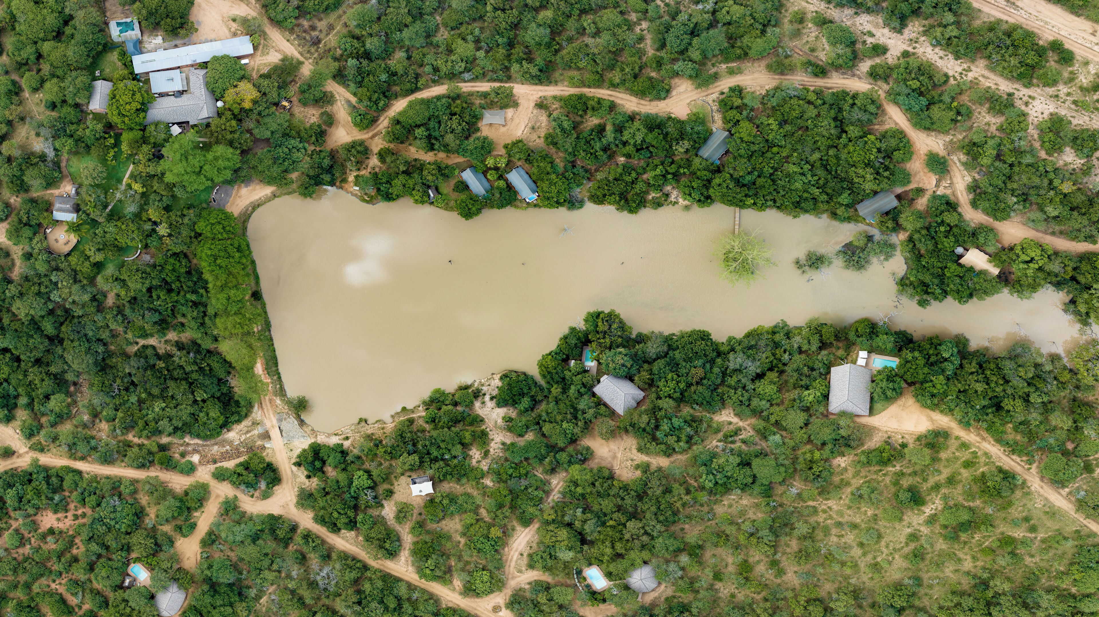 An aerial view of a property with a large water body, surrounded by dense greenery and several buildings with distinct roofs.