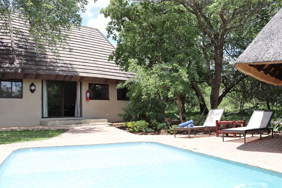 A pool area with lounge chairs, a house with a tiled roof, and a tree in the background.