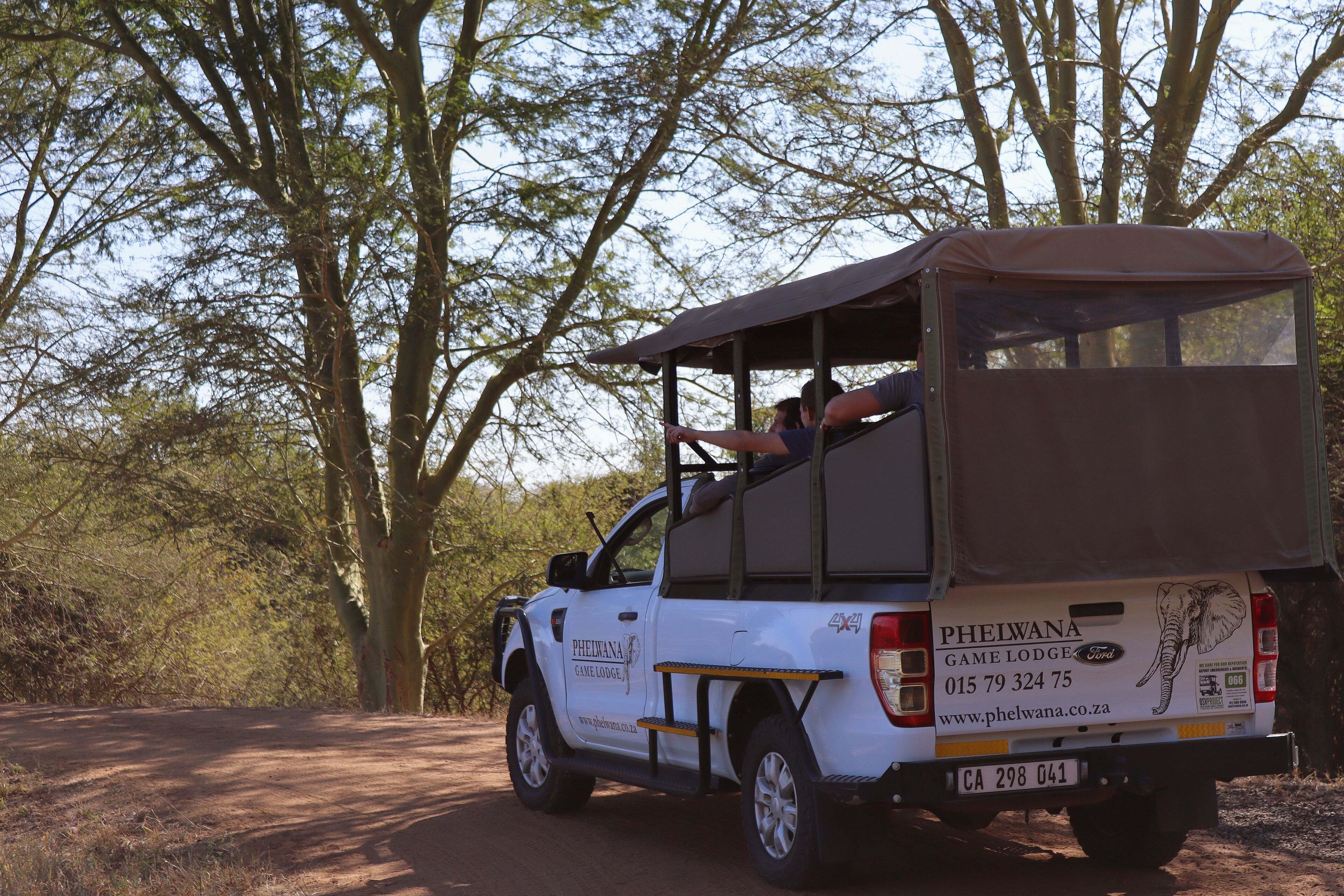 A white safari vehicle with a roof, parked on a dirt road surrounded by trees.