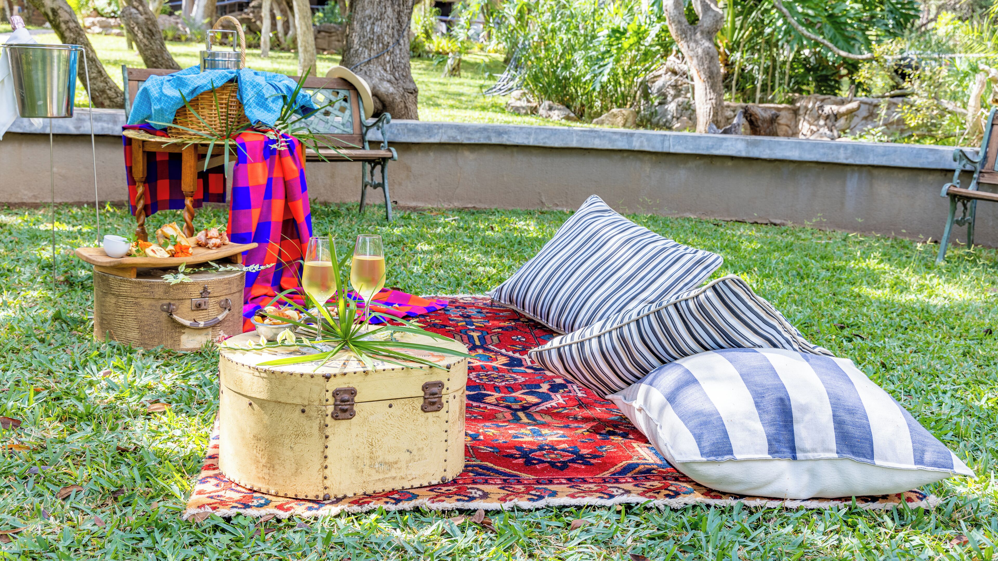 Montaje de picnic al aire libre con una cesta de picnic, una cesta tejida, un balde de metal, un caja de madera, un cojín a rayas y una manta estampada sobre una zona de césped.