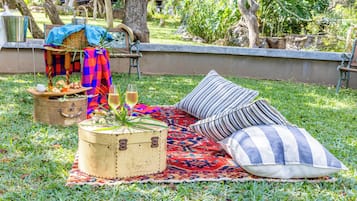 Montaje de picnic al aire libre con una cesta de picnic, una cesta tejida, un balde de metal, un caja de madera, un cojín a rayas y una manta estampada sobre una zona de césped.