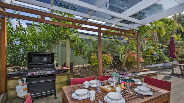 Een terras buiten met een houten eettafel, een barbecue en een pergola.
