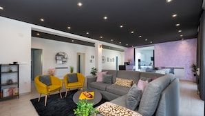 Un salon moderne avec un canapé gris, des fauteuils jaunes et une table basse. On aperçoit une cuisine à l’arrière-plan, avec un mur d’accent de couleur violette.