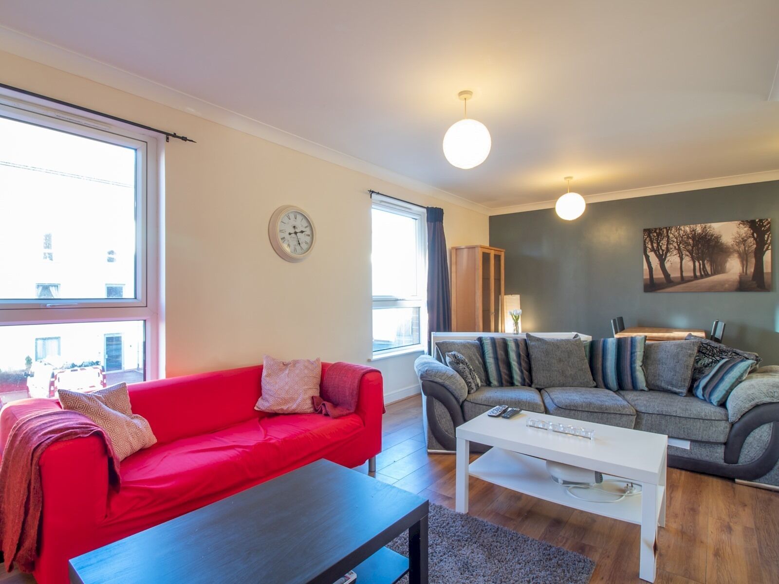 Una sala moderna con un sofá rojo, un sofá gris y una mesa de centro blanca. Hay un reloj en la pared y un cuadro sobre el sofá gris.