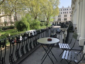 A balcony with a table and benches, overlooking a street with parked cars and buildings in the background.