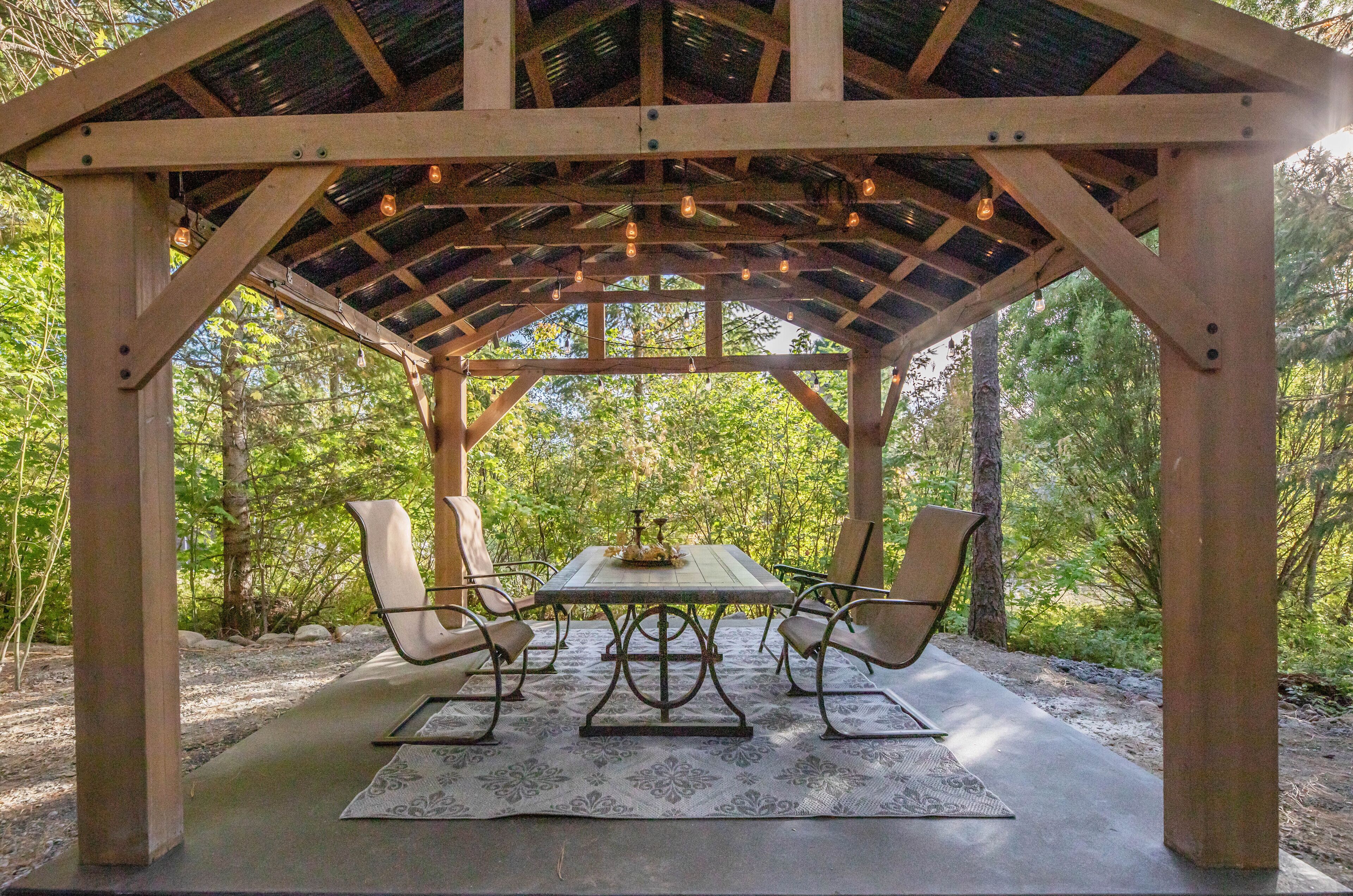 A wooden gazebo with a table and chairs, surrounded by trees.