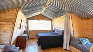 Un chalet confortable avec un toit en pente, des murs en bois, un lit au centre et un canapé agrémenté d’un coussin sur lequel est brodé le mot « bonjour ».
