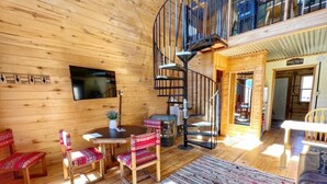 Un intérieur en bois avec un escalier en colimaçon, un coin repas avec des chaises, un téléviseur fixé au mur et une pancarte sur laquelle on peut lire « ENSEMBLE ».