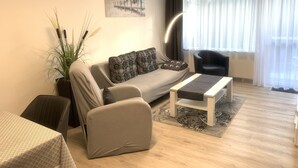 Una sala de estar moderna con un sofá, una mesa de centro y una silla. Hay una planta, un cuadro en la pared y una ventana con cortinas.